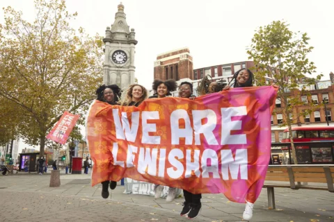 A group of smiling people jumping and holding a large banner reading "We Are Lewisham" in front of a clock tower. The scene is lively, with trees and buildings nearby.