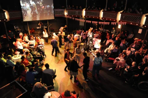 A lively ballroom scene in a warmly lit venue. Couples are dancing, surrounded by seated guests. Decorative bunting hangs above, creating a festive atmosphere.