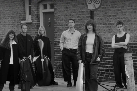 A black and white photo of a group of musicians stood on the street in front of a brick wall.