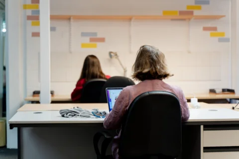 A photo of a woman with a bob working at a desk. Her back is facing the camera and she is working on a laptop with a pile of cables next to her.