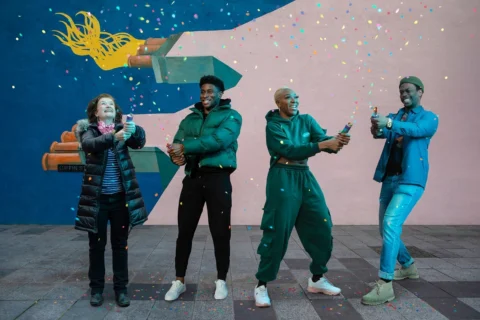 A photo of four people (Angela Clerkin, Wofai, DK, Ryan Calais Cameon) stood in front of a colourful mural in Deptford, holding confetti canons.