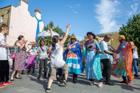 A group of people wearing colourful clothes stand in the middle of a square in Deptford on a sunny day.
