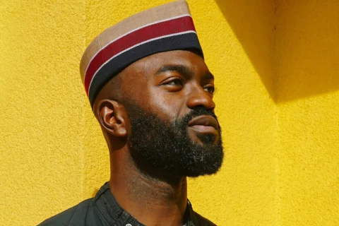 Inua Ellams - a black man wearing a red and black hat and a black jacket, stands in front of a bright yellow wall. He is gazing off to the right of the camera.