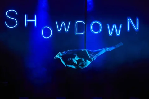 A photo of a performer suspended from a wire in the middle of a stage. Behind them the word 'Showdown' is illuminated in blue LED lighting.
