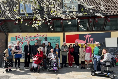 A photo of a group of older people (Meet Me at the Albany members) stood in front of a colourful mural on a sunny day. There are some branches with blossom on them at the top of the frame.