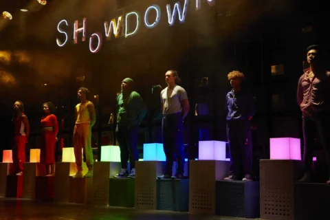 A photo of seven performers, each wearing a different bright colour and stood on a light up plinth in the same colour that they are wearing. Behind them the word 'Showdown' is lit in rainbow colours.