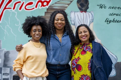 Albany Exec Director Mimi Findlay, Creative Director Vicki Amedume, and Chair of the Board Michelle Matherson, standing together in front of a colourful blue mural.