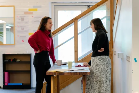 Two women with straight brown hair stand on either side of a low table, chatting and laughing.