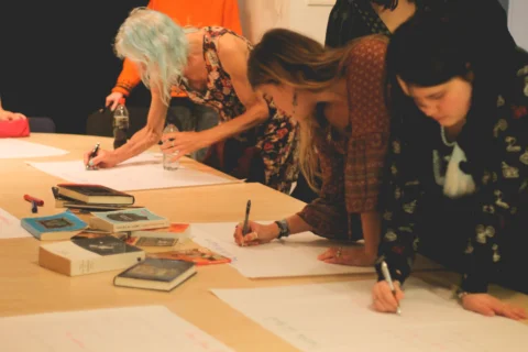 A photo of three women stood over a table, writing on large sheets of white paper. In the middle of the table are pens and a pile of books.