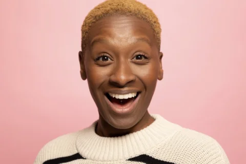A photo of comedian Chantel Nash - a black woman with short blond hair, wearing a black and white striped jumper. She is standing in front of a pink background and grinning widely at the camera.