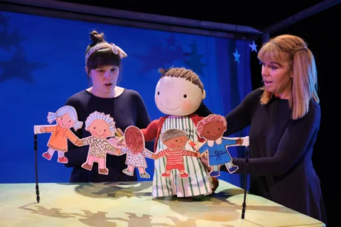 Two puppeteers dressed in black puppeteer a puppet of a young girl with brown hair, wearing a green and white striped dress and red cardigan. She is holding a string of paper dolls.