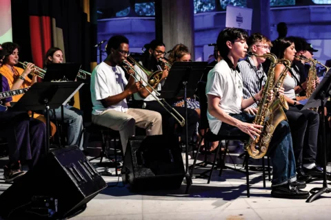 A photo of a brass band, with musicians playing the saxophone, trombone and trumpet, on a stage.