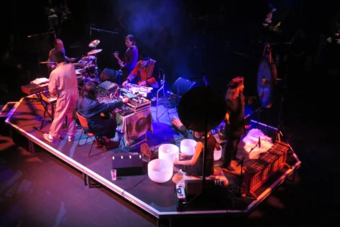 A photo of a stage from above. On the stage are musicians playing the drums, keyboards and guitar. They are lit in pink and purple lighting.
