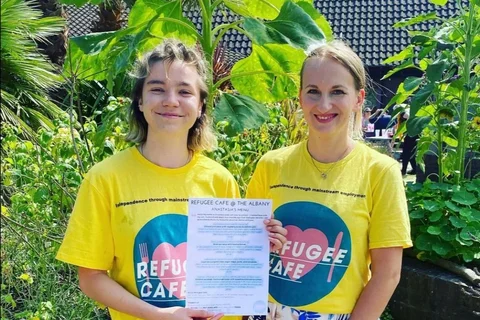 A photo of two white women wearing yellow t-shirts with blue and pink 'Refugee Cafe' logos on them. They are stood smiling in the Albany garden and are holding a cafe menu.
