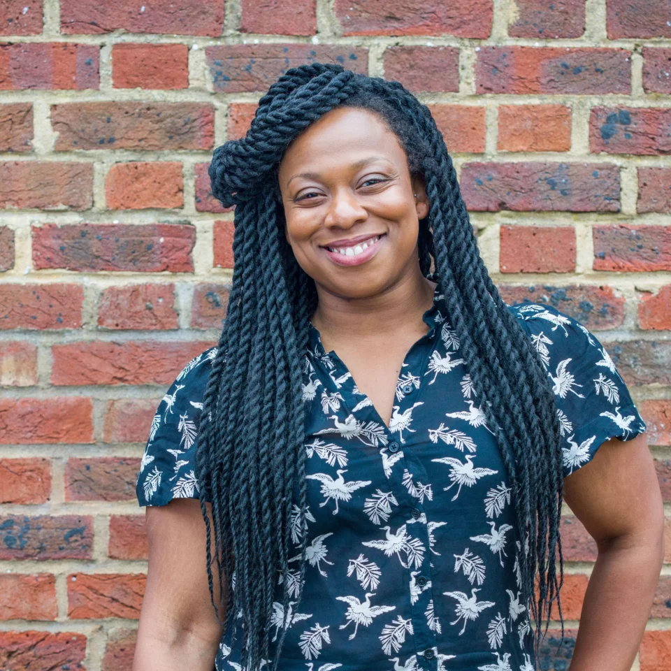 A photo of Vicki Amedume, a black woman with long braids, stood in front of a brick wall.