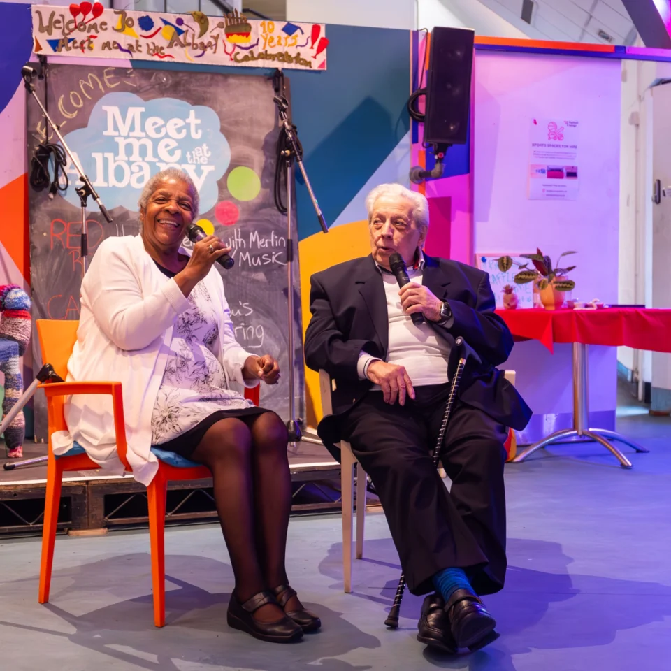 Elderly woman and man smiling, seated with microphones, against a colorful stage backdrop. Vibrant, inviting atmosphere with warm lighting.