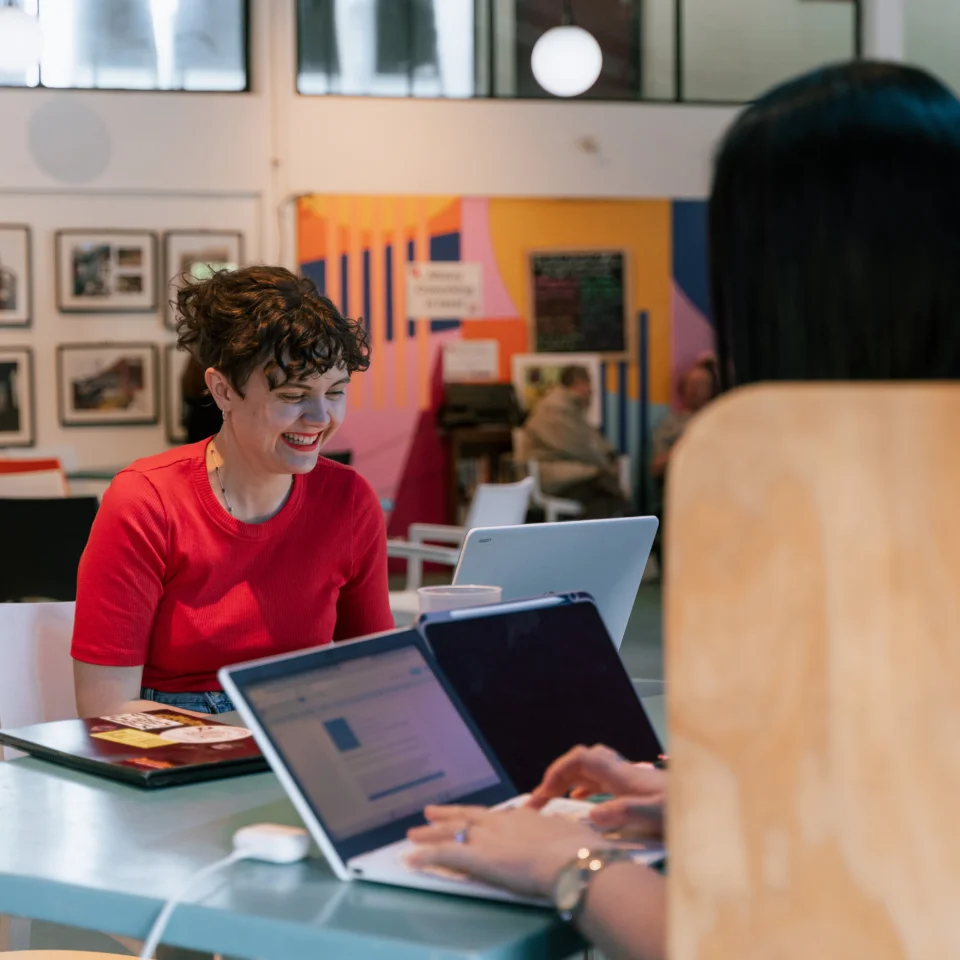 A photo of two people sat at a table on laptops. One has their back to the camera. The other is a white woman with a red top and curly hair, she is smiling.